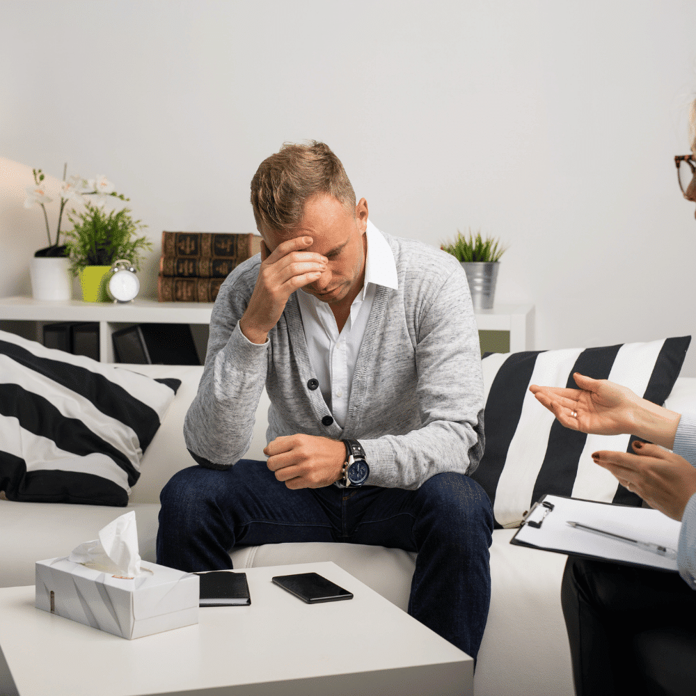 A man sits on a couch with his head in his hand, appearing stressed or upset. A tissue box and phone are on the table in front of him, while another person sits nearby, holding a clipboard and gesturing.