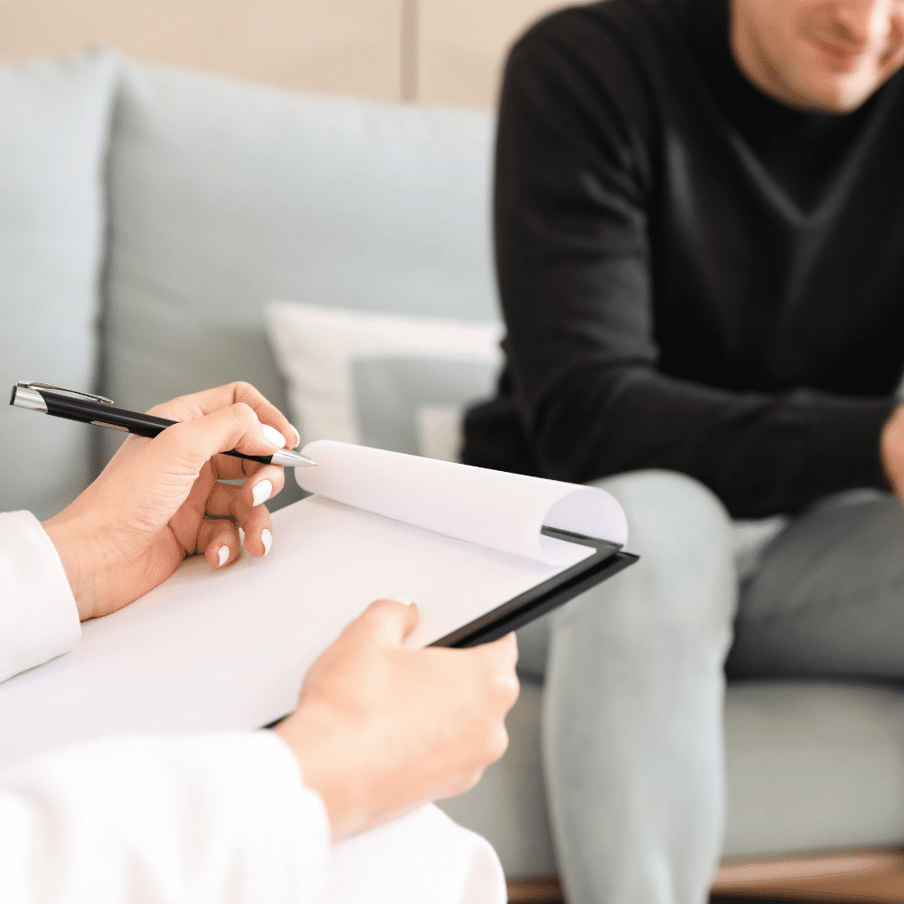 A person holding a pen and clipboard takes notes while another person sits on a couch, suggesting a therapy or counseling session in a comfortable indoor setting.