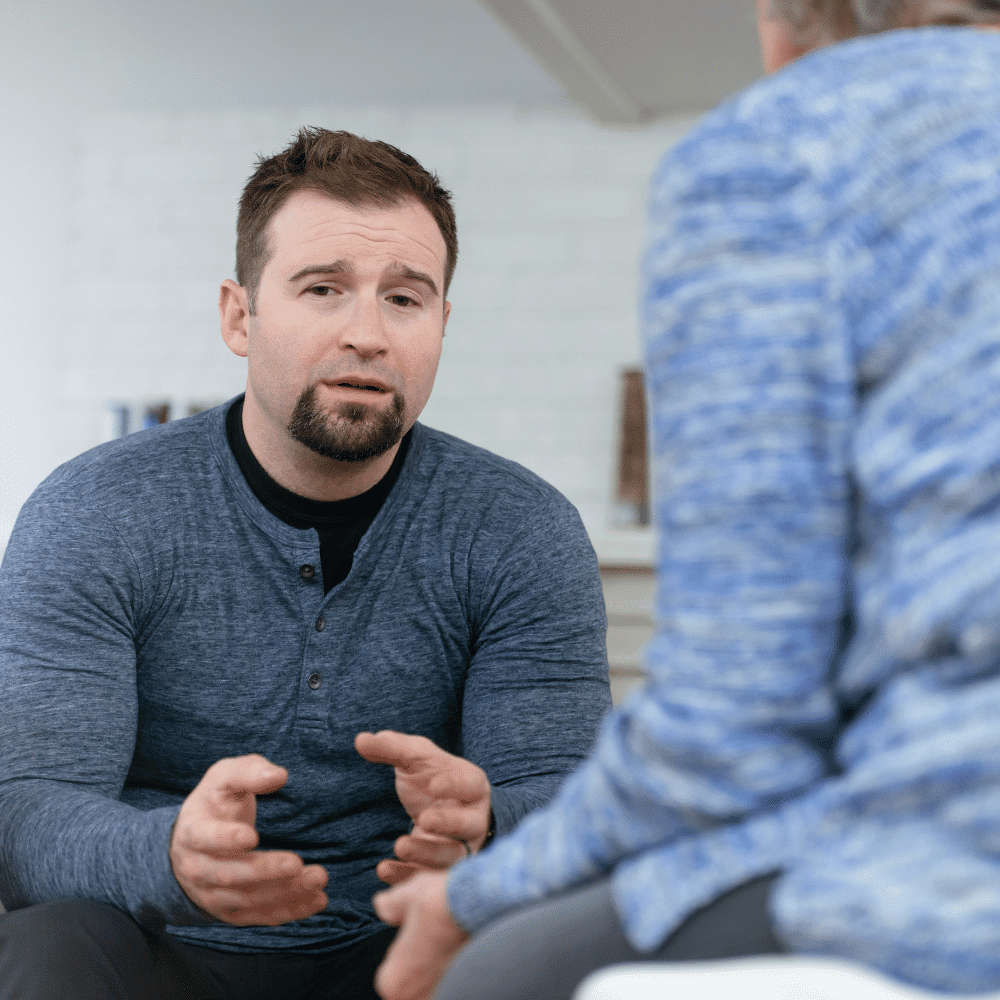 A man with short brown hair and a beard, wearing a blue shirt, sits indoors and gestures with his hands while talking to another person in a blue sweater, whose back is facing the camera.