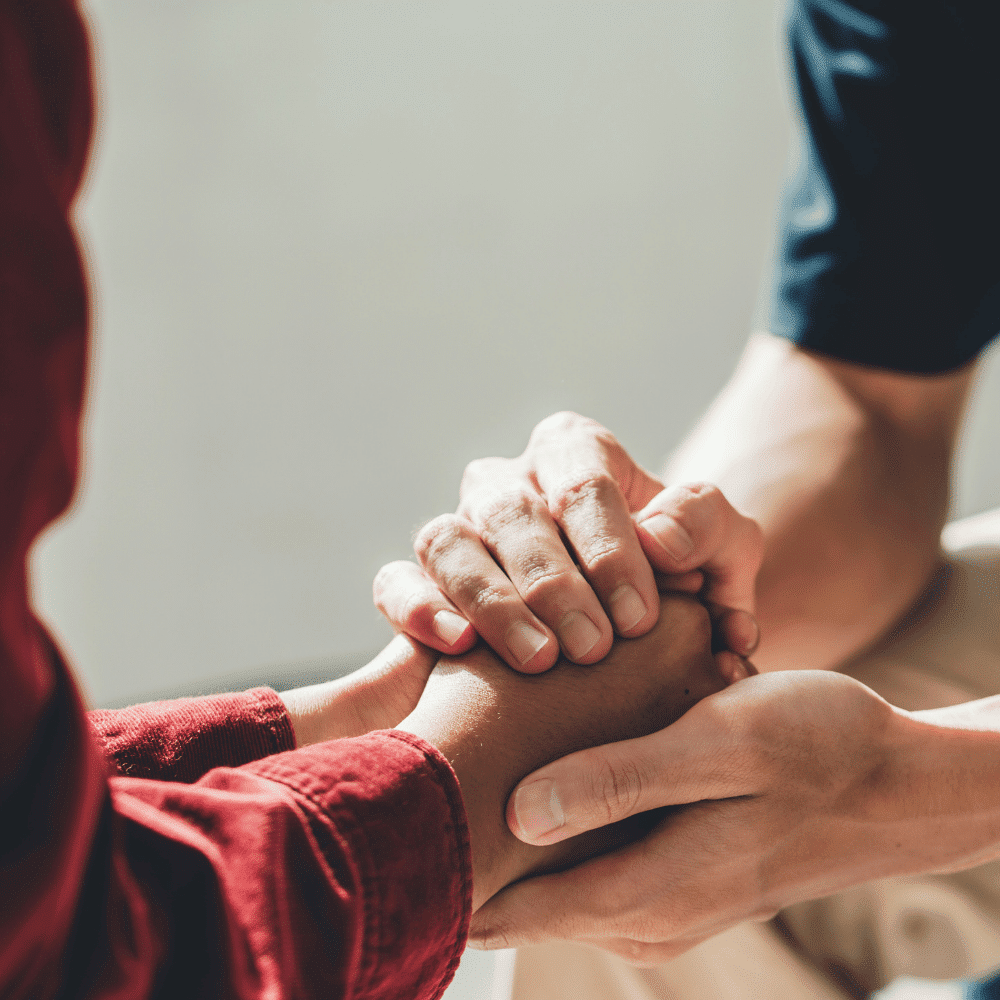 Close-up of two people holding hands in a comforting gesture. One person wears a red long-sleeve shirt, while the other is in short sleeves, offering support and empathy.