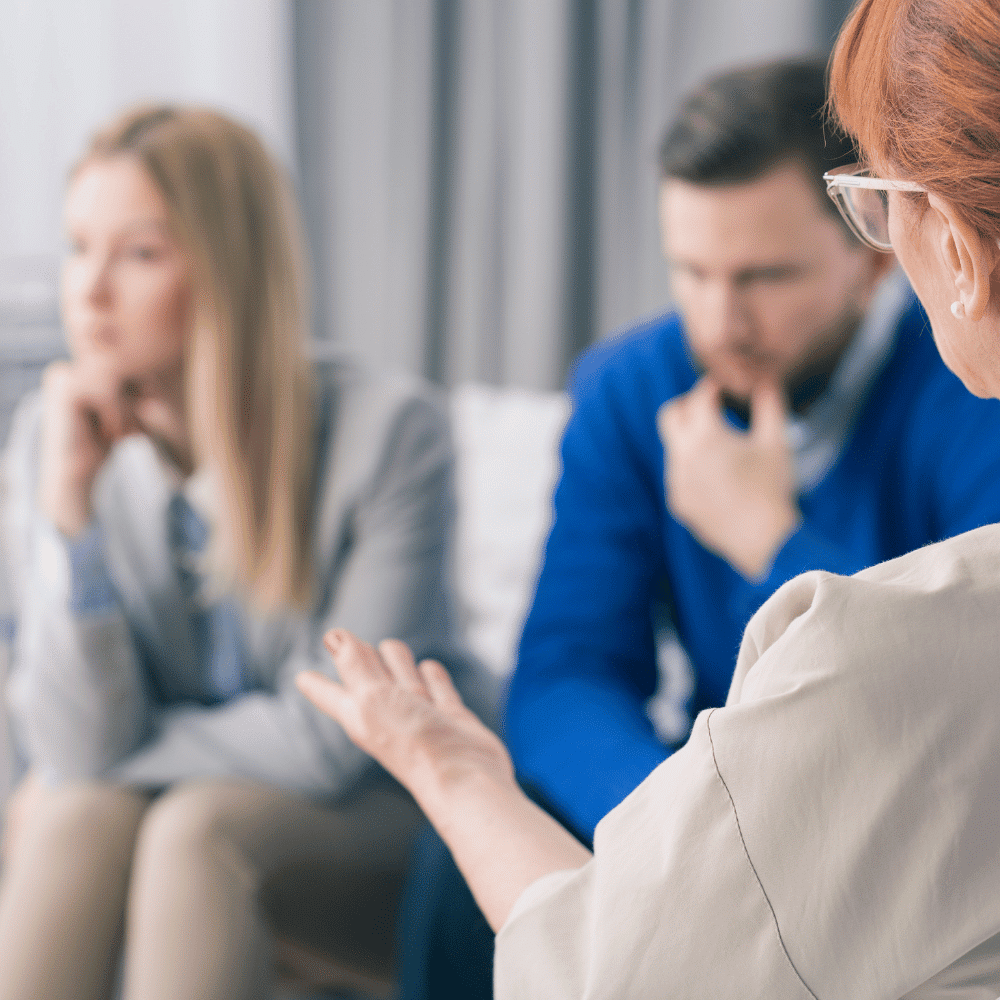 Three people sit together indoors, appearing engaged in a serious conversation. One person gestures while speaking, while the other two listen attentively, with blurred backgrounds emphasizing their focus.