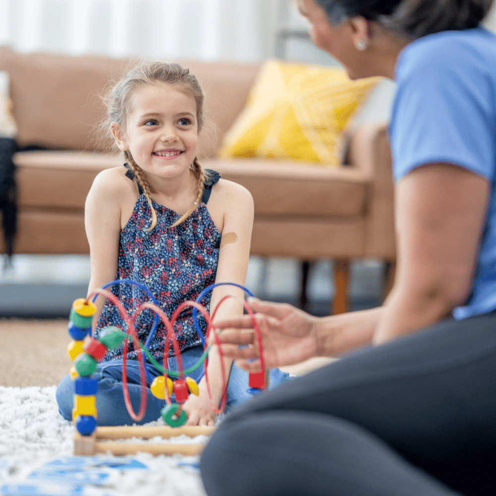 A young girl smiles while playing with a colorful bead maze toy on the floor, sitting across from an adult in a bright living room with a sofa and yellow pillow in the background.